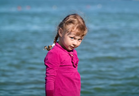 little girl with blue eyes in a raspberry dress looking slyly at the background of waterの写真素材