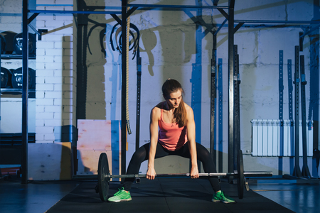 Muscular young fitness woman lifting a weight crossfit in the gym. Fitness woman deadlift barbell. The gym on the red wall is written SPORT. Crossfit woman. Crossfit style.の写真素材