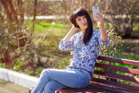 Beautiful asian girl sitting on bench in park, making selfie on smartphone