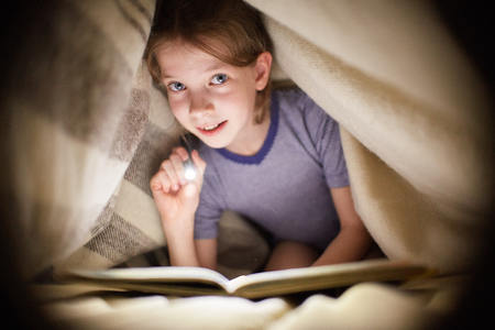 little girl is reading a book under a blanket with a flashlight in a dark room at nightの写真素材