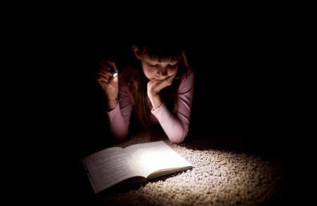 little girl is reading a book with a flashlight in a dark room at nightの写真素材