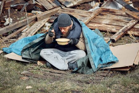 a homeless man eats soup from a plate near the ruins, helping poor and hungry people during the epidemicの写真素材