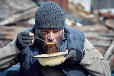 a homeless man eats soup from a plate near the ruins, helping poor and hungry people during the epidemicの写真素材