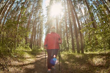 a woman in a protective mask walks in a Park with sticks for Nordic walking, a walk in the fresh air after quarantine, a precautionary measure against the virusの写真素材