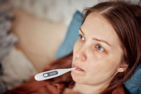 high temperature - the first sign of a viral disease, a woman measures the temperature with a thermometer.の写真素材