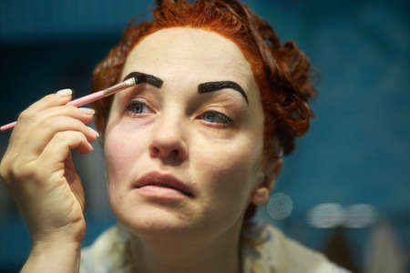 red-haired forty-year-old woman paints her eyebrows with paint in the bathの写真素材