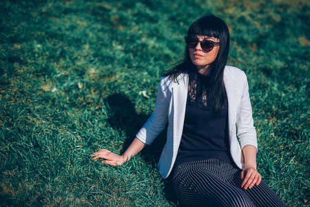 portrait of a life style brunette girl in sunglasses on the grass in the park in a white jacket