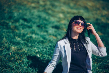 portrait of a life style brunette girl in sunglasses on the grass in the park in a white jacketの写真素材