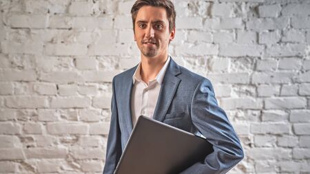Photo a positive Manager holding a laptop and smiling, against a white brick wall. close upの写真素材