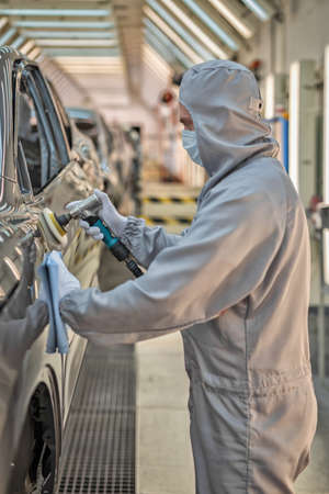 An employee of the car body paint shop in white gloves and a medical mask on his face polishes the painted surface. Work during the pandemic. Automobile plantの写真素材