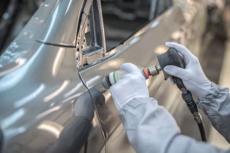 An employee of the car body paint shop in white gloves and a medical mask on his face grinds the...
