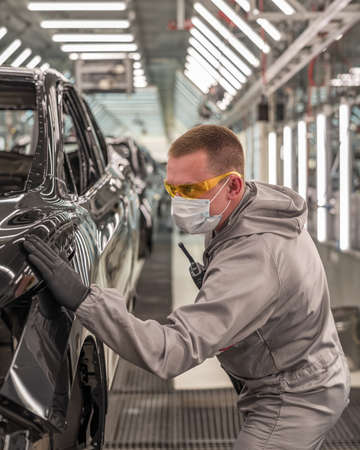 An employee of the paint shop of an automobile factory in a medical mask on his face checks the...