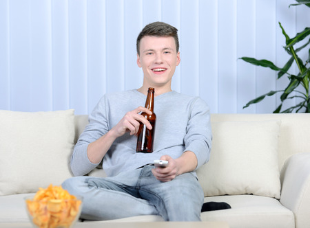 Relaxed young man watching tv and drinking beer at home in bright living roomの写真素材