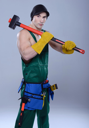 Handsome young topless construction worker with sledge hammer, studio shot, gray backgroundの写真素材