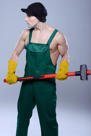 Handsome young topless construction worker with sledge hammer, studio shot, gray backgroundの写真素材