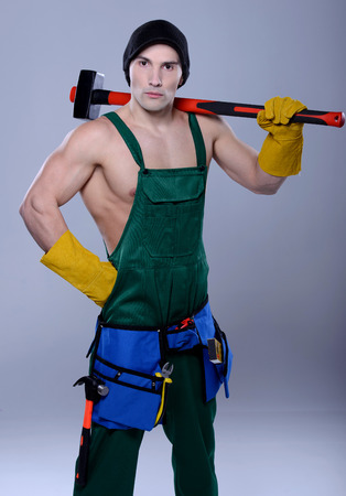 Handsome young topless construction worker with sledge hammer, studio shot, gray backgroundの写真素材