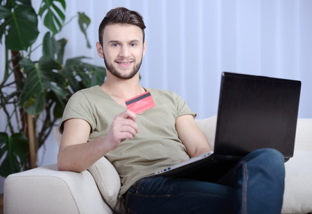 Cheerful handsome man in bright living room using his credit card to buy onlineの写真素材