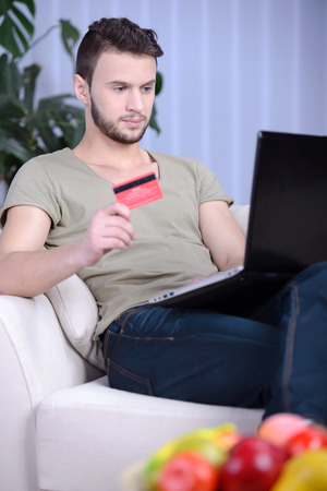 Cheerful handsome man in bright living room using his credit card to buy onlineの写真素材
