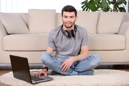 Man listening to music with earphones on laptop computer, sitting on the floor near the sofaの写真素材