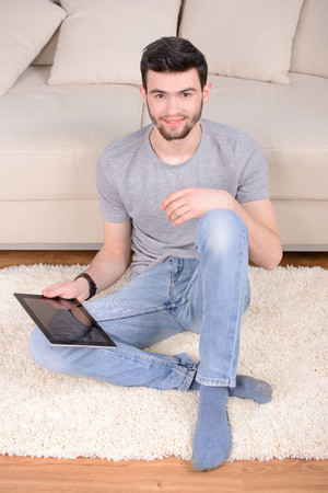Handsome casual young man using a tablet at home sitting on the floor next to a sofa in his living room browsing the internet with a smileの写真素材