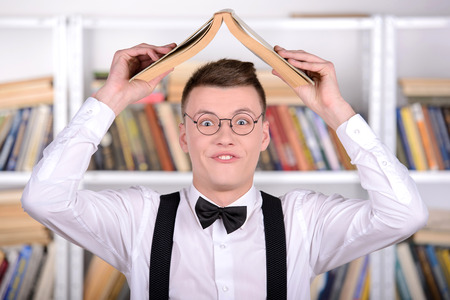 Smart young man intelehent in shirt and tie tie and glasses holding book on head standing in the libraryの写真素材