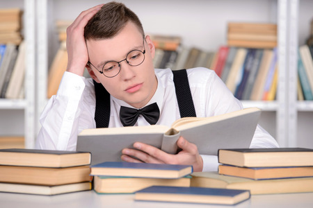 Smart young man intelehent in shirt and tie tie and glasses reading a book on the head while standing in a libraryの写真素材