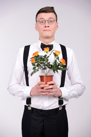 Young botanist. Young man in bow tie and suspenders holding a plant in his hands and smiling while standing against grey backgroundの写真素材