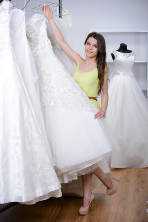 Smiling pretty bride chooses white gown at shop of wedding fashionの写真素材