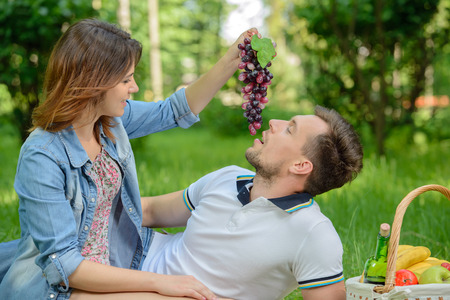 Lovely moments. Young girl feeding her boyfriend with a grapes bunch on the picnicの写真素材