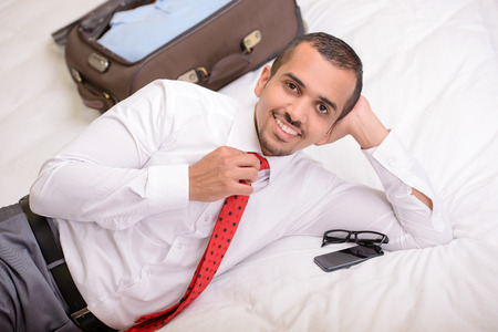 Portrait of Asian businessman with a suitcase lying on the bed in the hotel roomの写真素材