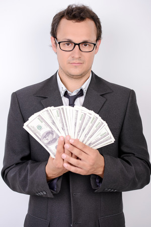 Cheerful young man in formalwear and glasses holding paper currency and smiling at camera while standing against grey backgroundの写真素材