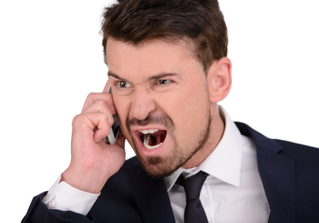 Angry businessman with mobile phone. Furious young businessman holding a mobile phone and pointing it while isolated on whiteの写真素材