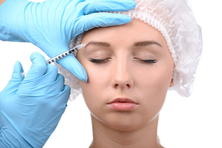 Medical injection. Portrait of beautiful young woman in medical headwear looking away while hand in glove making an injection in her face isolated on whiteの写真素材