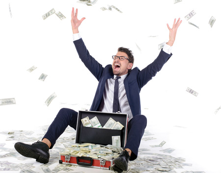 I am rich! Happy young businessman in formalwear throwing money up while sitting near the case full of paper currencyの写真素材