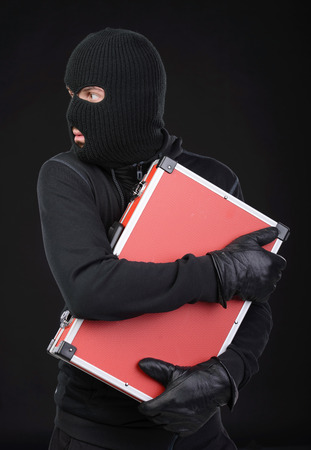 Stealing a suitcase. Frustrated thief in black balaclava holding a stolen suitcase while isolated on blackの写真素材
