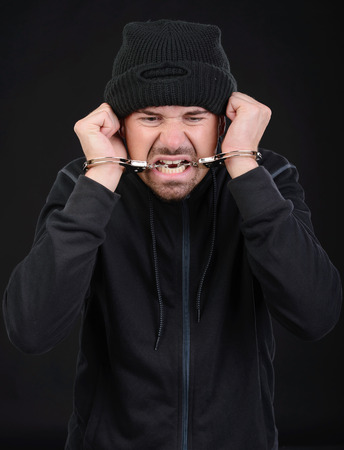 Busted burglar. Angry burglar in handcuffs grimacing at camera while standing against black の写真素材