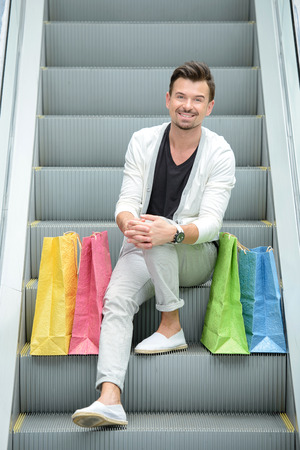 Young handsome man with bags for shopping, standing on escalator in shopping centerの写真素材