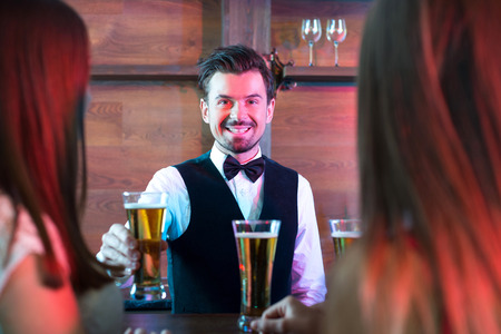 Freshly tapped beer. Handsome smiley bartender stretching out beer mugs and smilingの写真素材