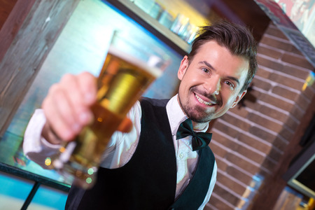 Freshly tapped beer. Handsome smiley bartender stretching out beer mugs and smilingの写真素材