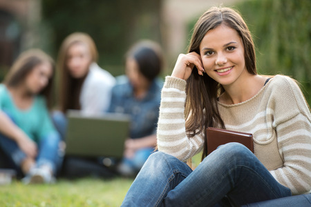 Portrait of a beautiful, smiling college girl is holding book with blurred students are sitting in the park.の写真素材