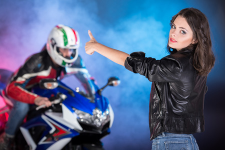 Portrait of young attractive woman. Biker on a motorcycle behind her on colored, foggy background.
