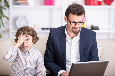 A young father is working at the computer. Son sits beside him.の写真素材
