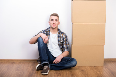 Young man is sitting on the floor next to cardboard boxes. Moving into a new house.