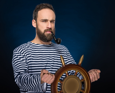 Young bearded sailor in striped clothing is smoking a pipe while standing with steering wheel.