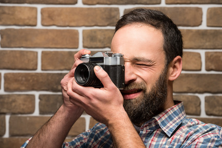 Young bearded man with vintage camera standing against brick wall.