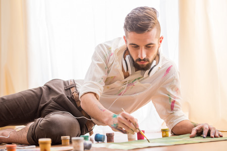 Young artist is painting on canvas is lying on studio floor while listening a music.