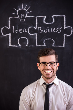 Handsome young man is standing against blackboard with connecting jigsaw pieces. Idea and business concept.の写真素材