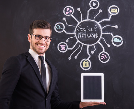 Social networking. Smiling young man is holding tablet while standing against social network chalk drawing on blackboard.の写真素材