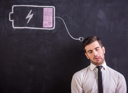 Young tired man is standing against the blackboard with drawing battery low.の写真素材