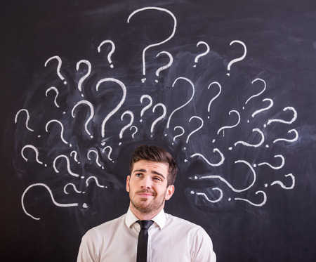 Young man is standing against blackboard with question marks.の写真素材